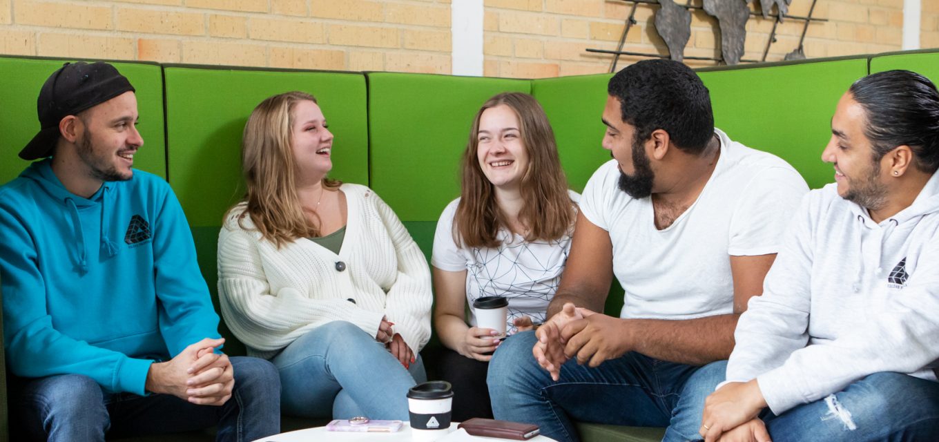 Fem deltagare allmän kurs umgås i grön soffa, kaffekollar och hoodies har Eslövs folkhögskolas logotyp