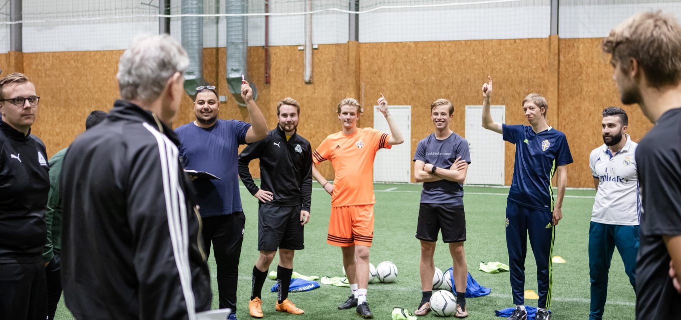 Deltagare på fotbollstränarlinjen står i en ring, tre med handen uppräckt, vända mot läraren i konstgräshallen