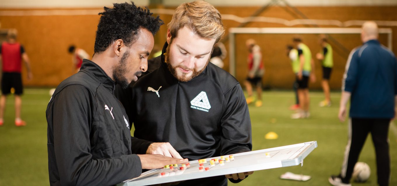 Två deltagare på fotbollstränarlinjen diskuterar en taktiktavla med klassen i bakgrunden i konstgräshallen