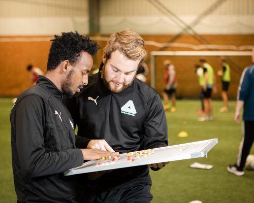 Två deltagare på fotbollstränarlinjen diskuterar en taktiktavla med klassen i bakgrunden i konstgräshallen