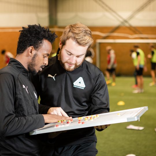 Två deltagare på fotbollstränarlinjen diskuterar en taktiktavla med klassen i bakgrunden i konstgräshallen