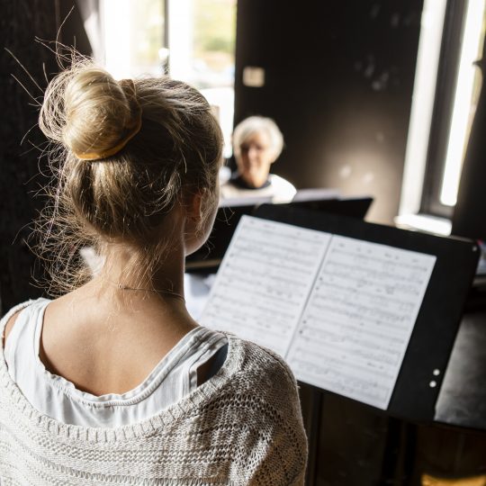 En deltagare vid ett notställ vänd från kameran emot sånglärare vid flygel i bakgrunden