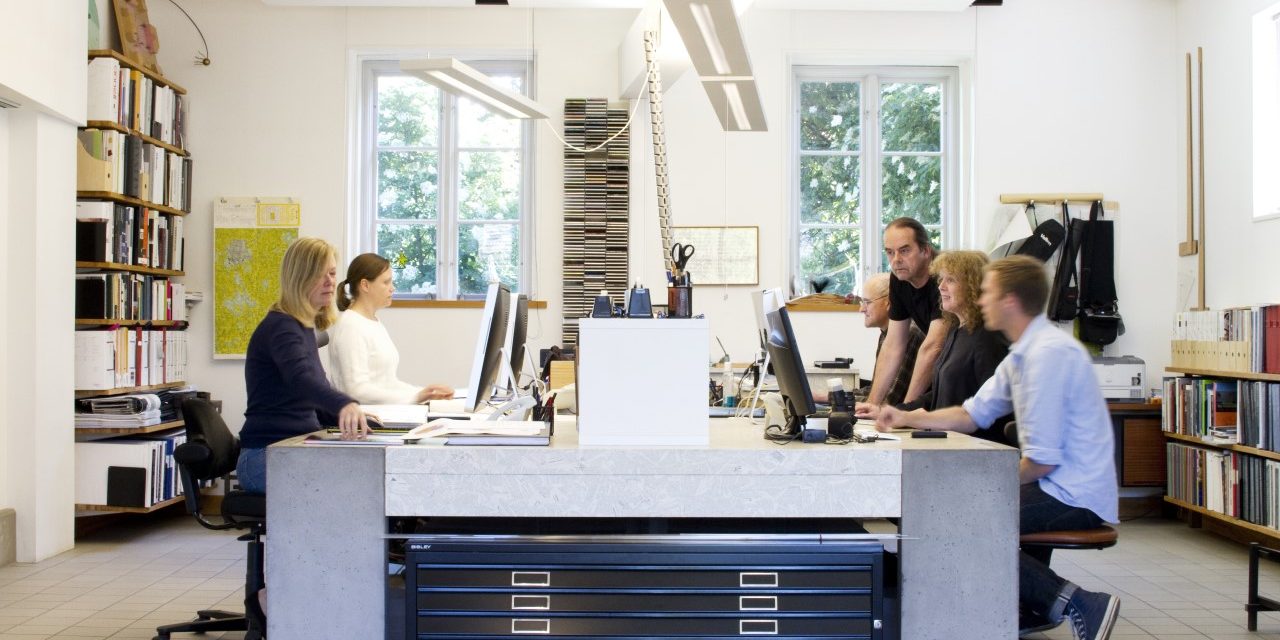 En bild från Lokal XXX arkitekters kontor med sex personer arbetandes runt ett gemensamt bord med datorer och kontorsmaterial