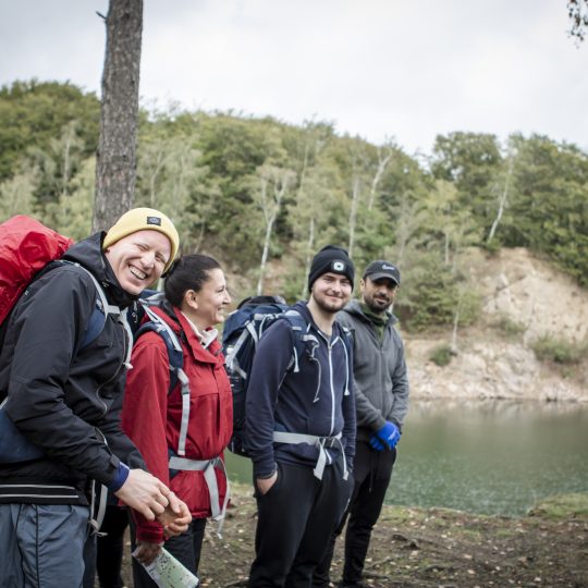 Fyra deltagare fritidsledarutbildningen på naturvandring med ryggsäckar intill en sjö med skog i bakgrunden